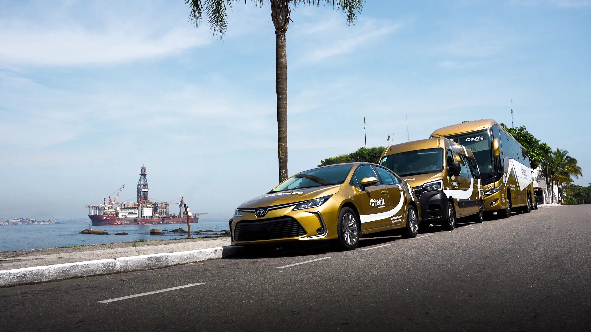 Corolla, van e ônibus da frota Braslectra na orla, com plataforma offshore ao fundo